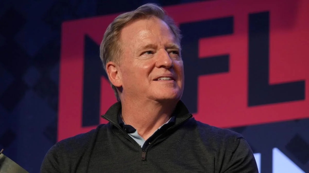 Roger Goodell speaks onstage in a dark pullover before a large NFL backdrop.