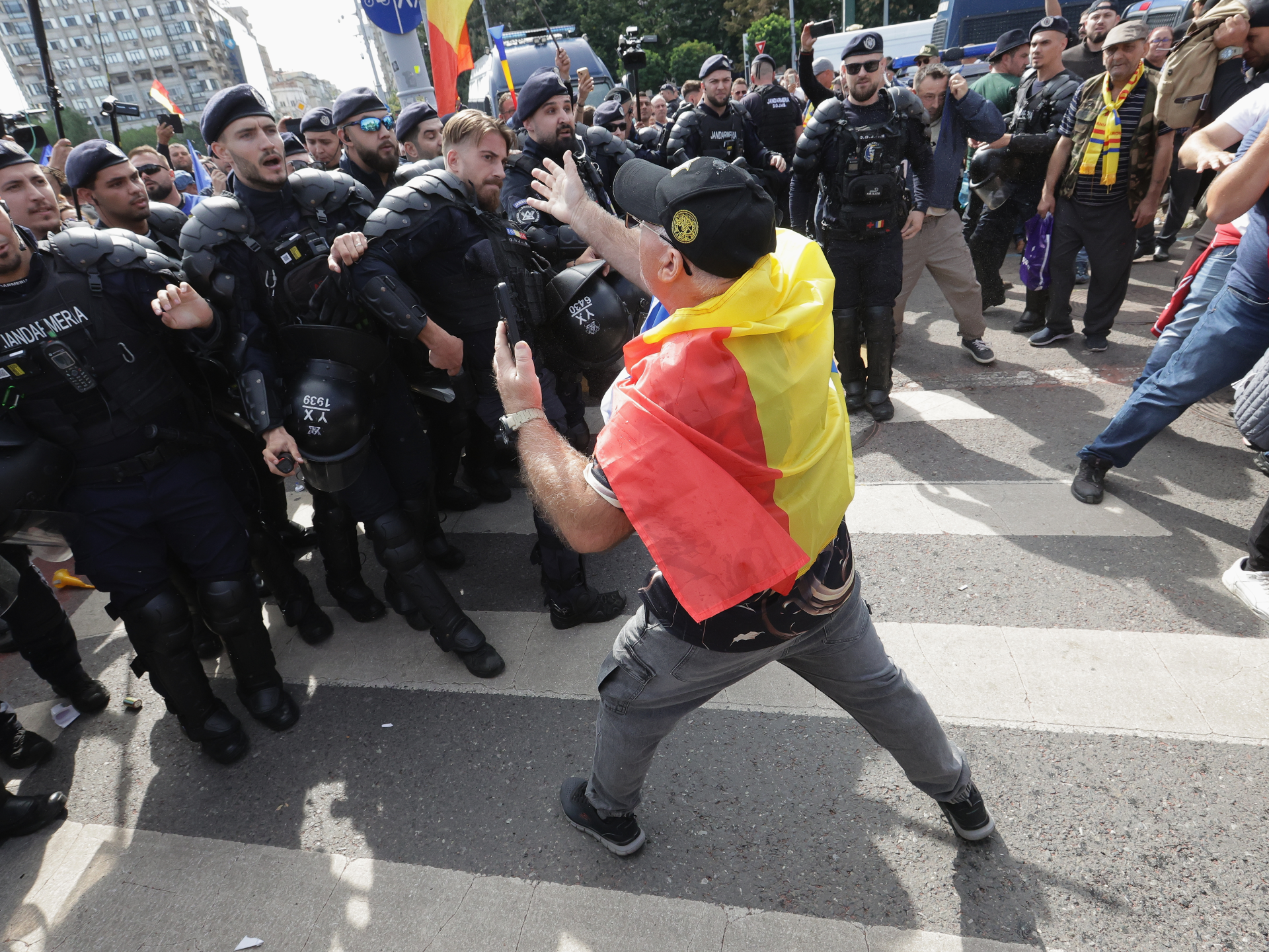 Man draped in Romanian flag gestures toward a line of riot police during a street confrontation