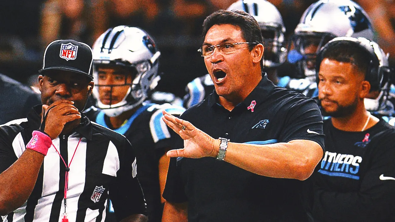 Football coach gestures on the sideline as players and staff watch during a game.