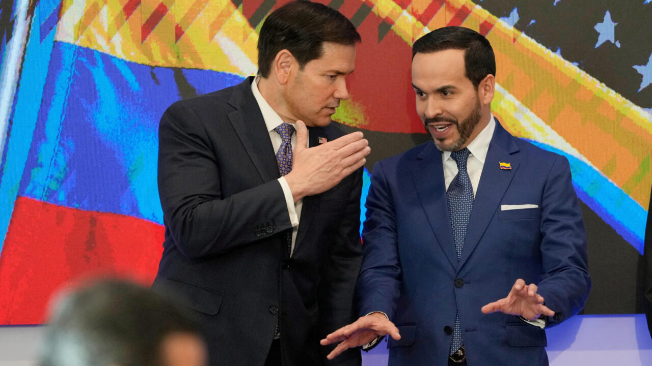 Two suited men converse and gesture before a colorful Colombian flag backdrop.