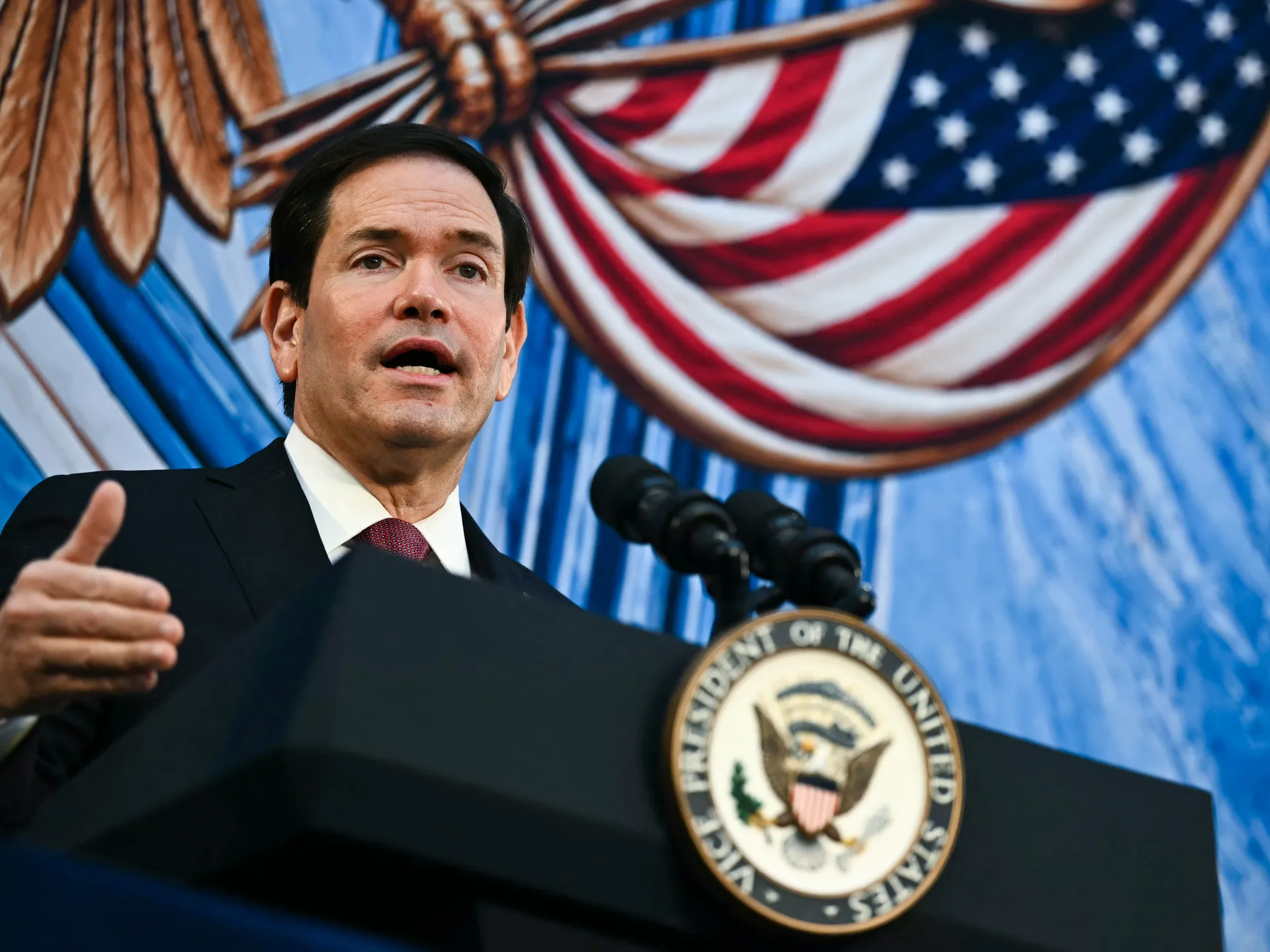 Man speaking at a podium with microphones, U.S. flags and patriotic bunting behind him