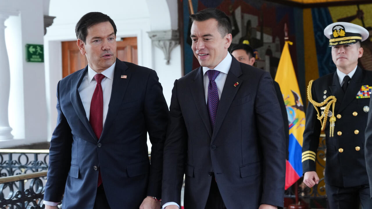 Two suited officials walk indoors beside an Ecuadorian flag and a uniformed naval officer.