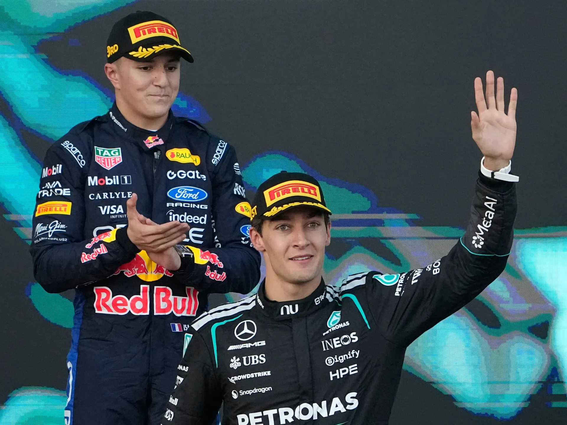 A Mercedes driver waves from the podium while a Red Bull driver stands behind him clapping.