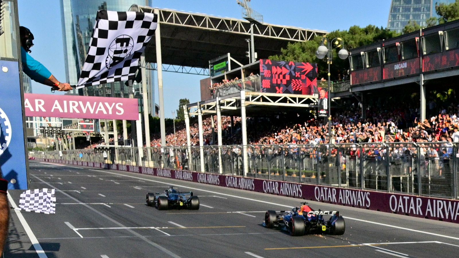 Two Formula One cars race down the straight as a marshal waves a checkered flag beside the track.
