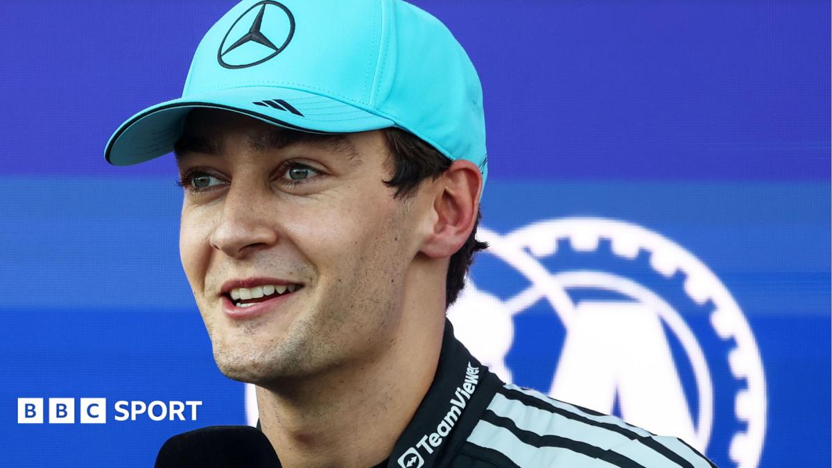A smiling man in a turquoise cap and dark team jacket poses against a blue background with a large white emblem.