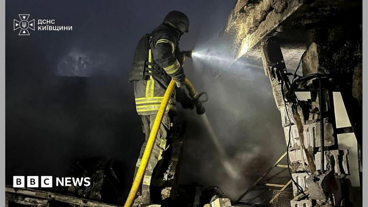 A firefighter sprays water onto smoke-filled rubble beside a severely damaged structure at night.