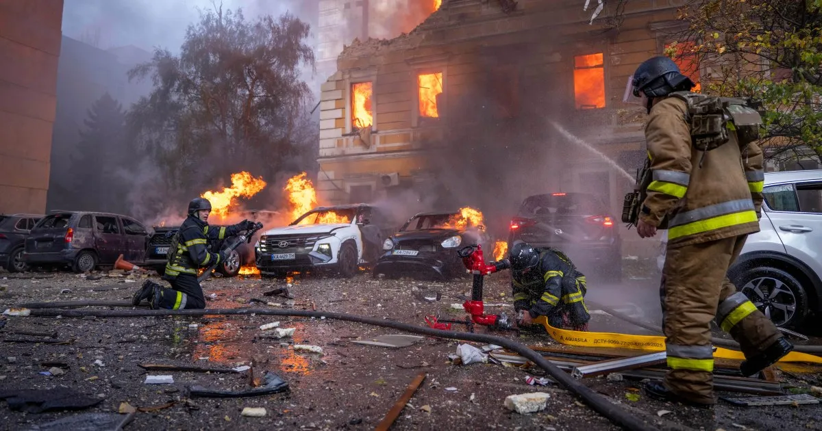 Firefighters tackle burning cars beside a smoke-filled building as flames glow through its windows.