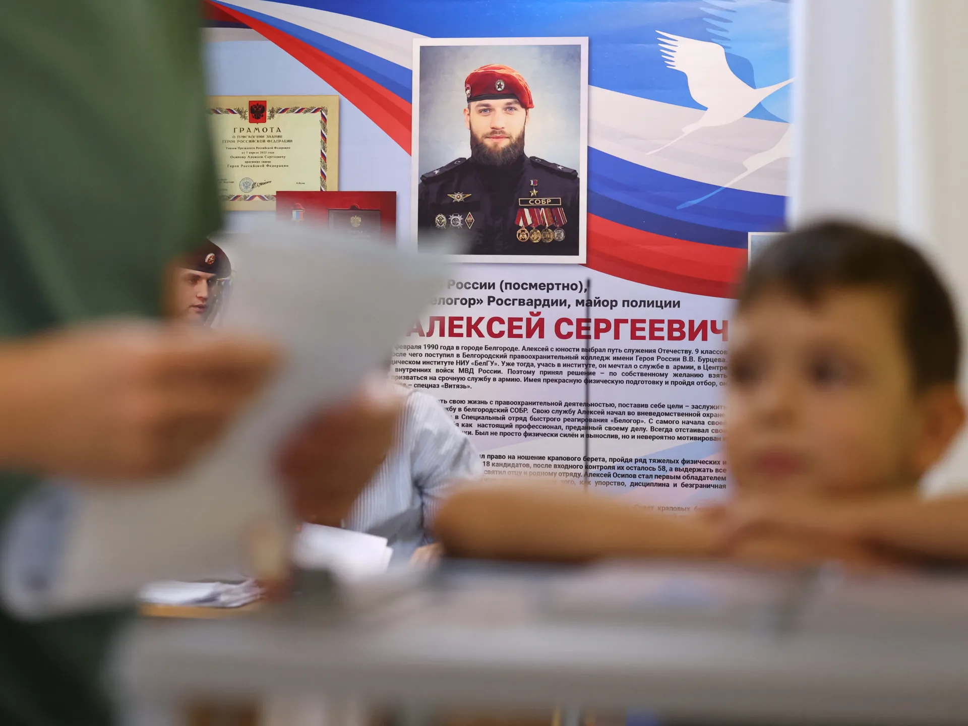 Blurred people sit before a memorial display featuring a uniformed man’s portrait and Russian flag imagery.