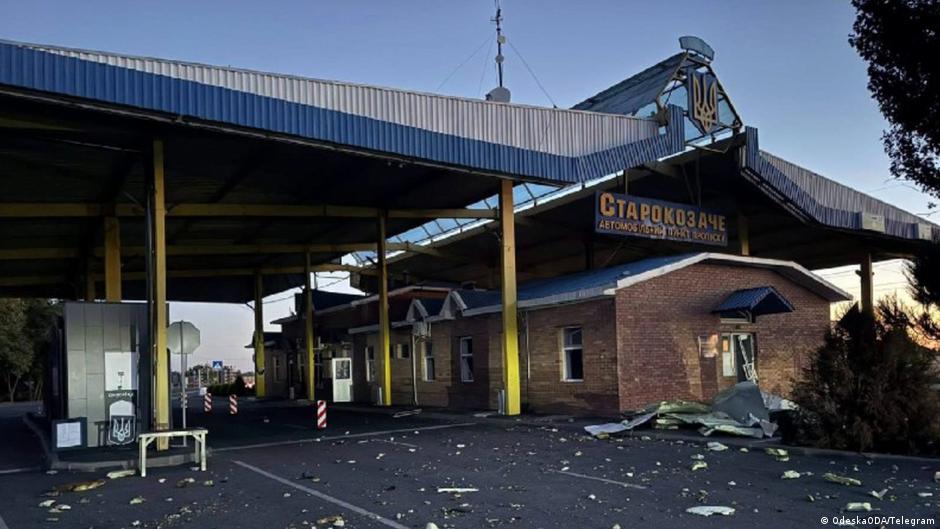 Damaged roadside fuel station with shattered windows, debris, and torn roofing beneath a large canopy.