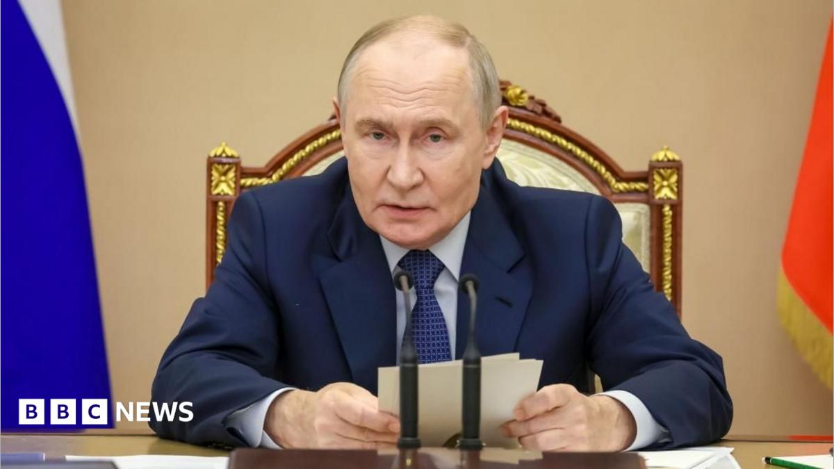 Russian president seated at a desk between microphones, holding papers in front of national flags