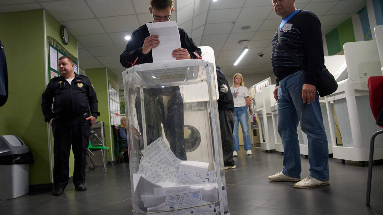 People stand around a transparent ballot box as an election official reads a document indoors.