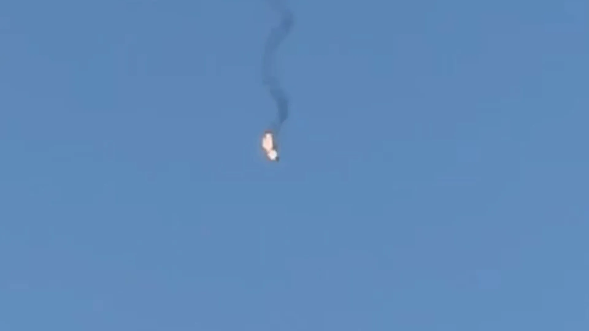 A small bright object descends through a clear blue sky, trailing a winding plume of dark smoke.