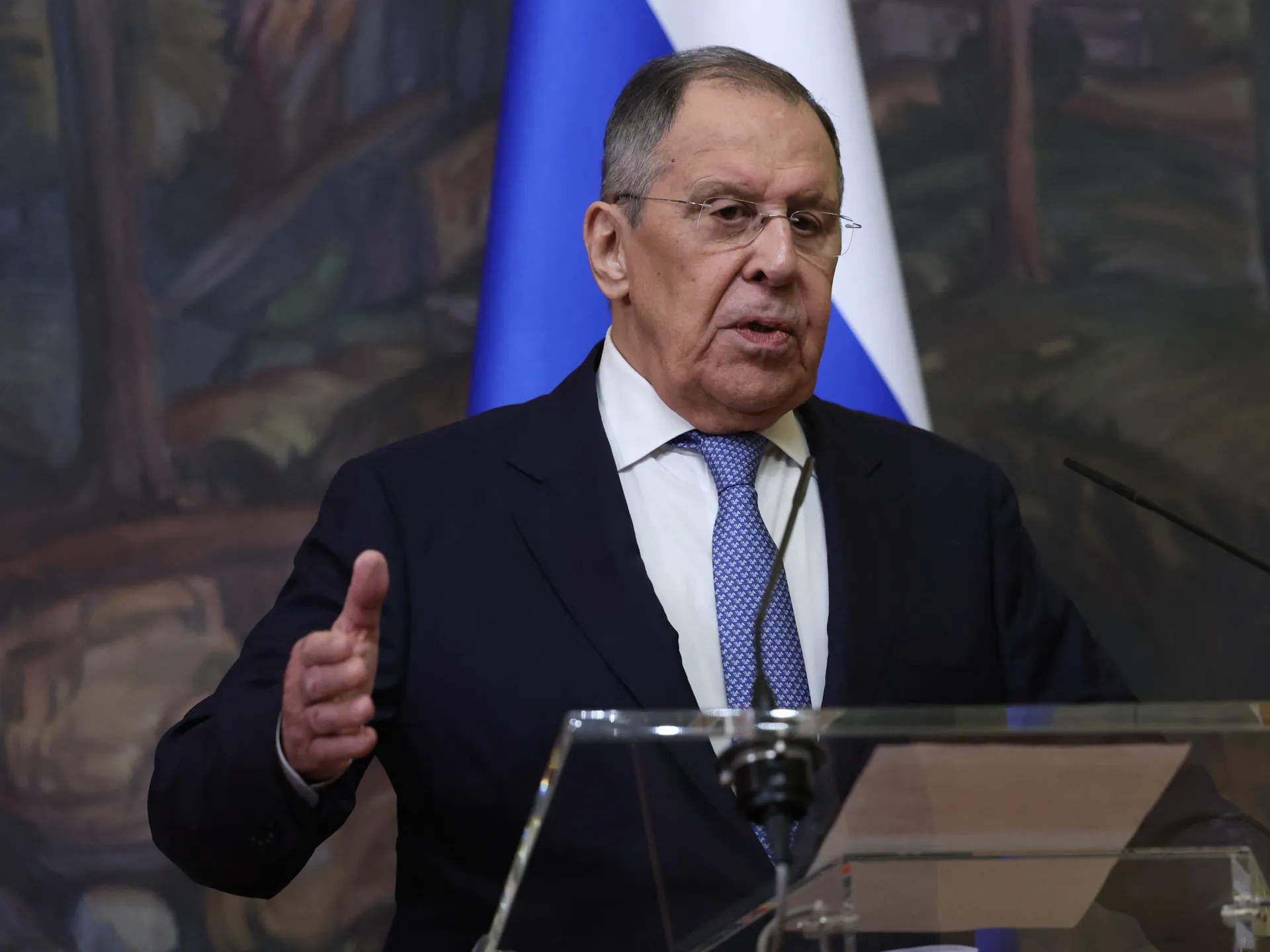 Man in suit and glasses speaks at a lectern with a Russian flag behind him