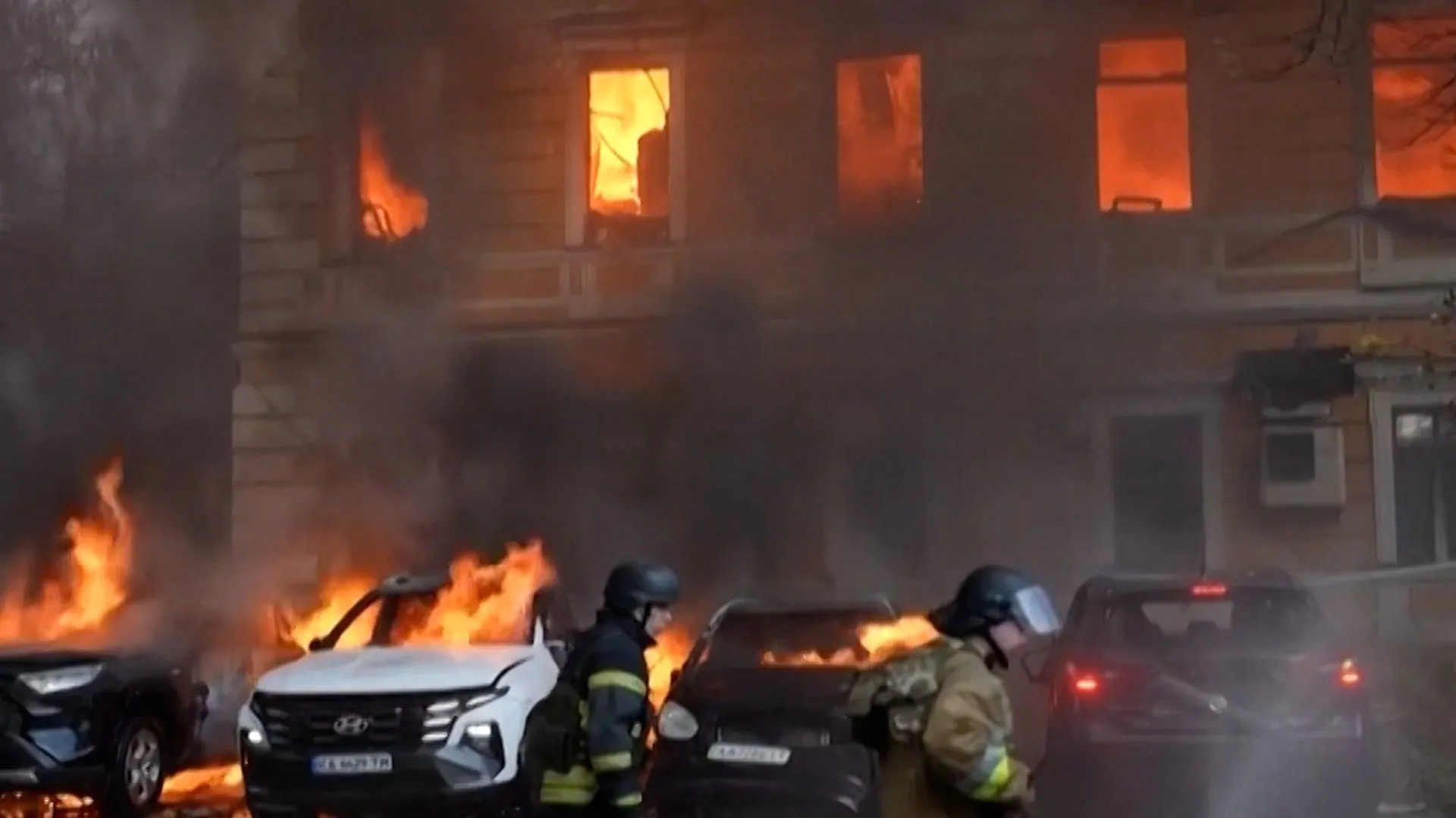 Firefighters stand beside burning cars as flames glow through windows of a smoke-filled building.