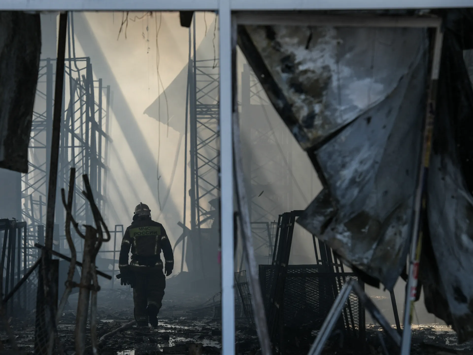 Firefighter walks through a smoke-filled, heavily damaged industrial structure with twisted metal and debris.