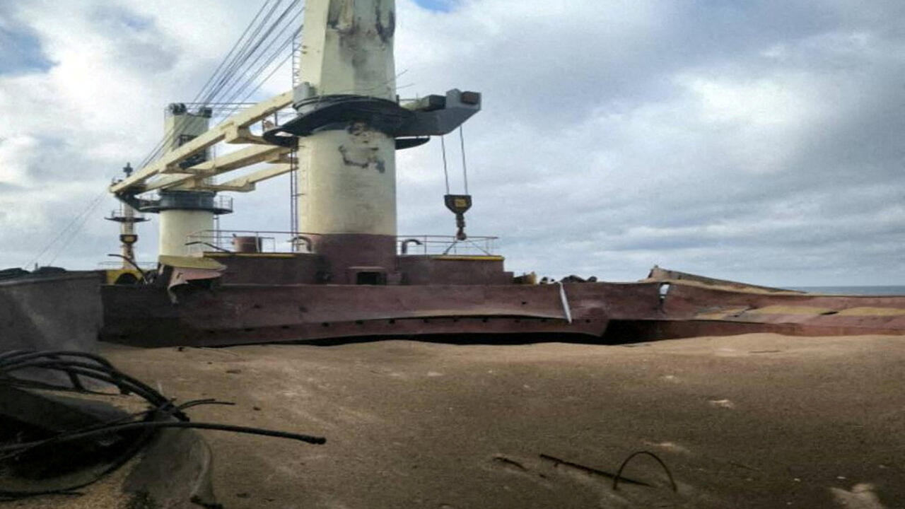 A rust-colored cargo vessel sits grounded beside a concrete pier beneath cloudy skies.