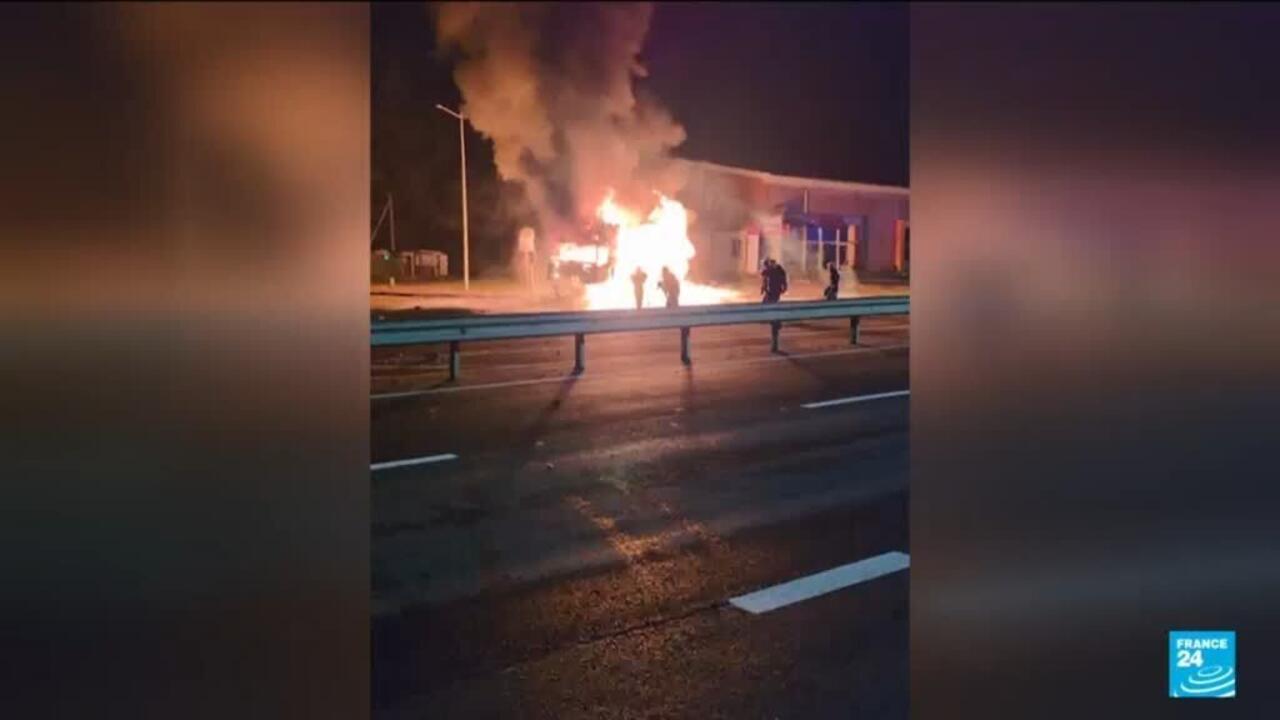 Large fire and thick smoke rise beside a roadside building as silhouetted figures stand nearby.
