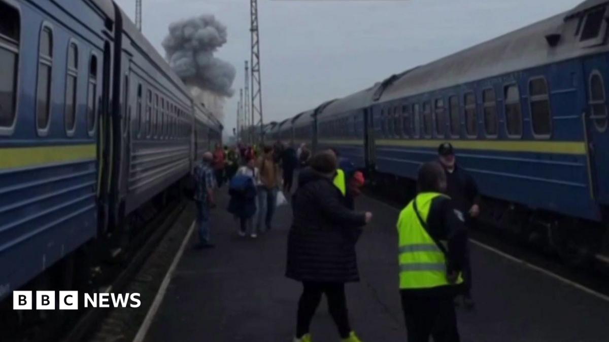 People stand between blue-and-yellow passenger trains as a large smoke plume rises behind them.