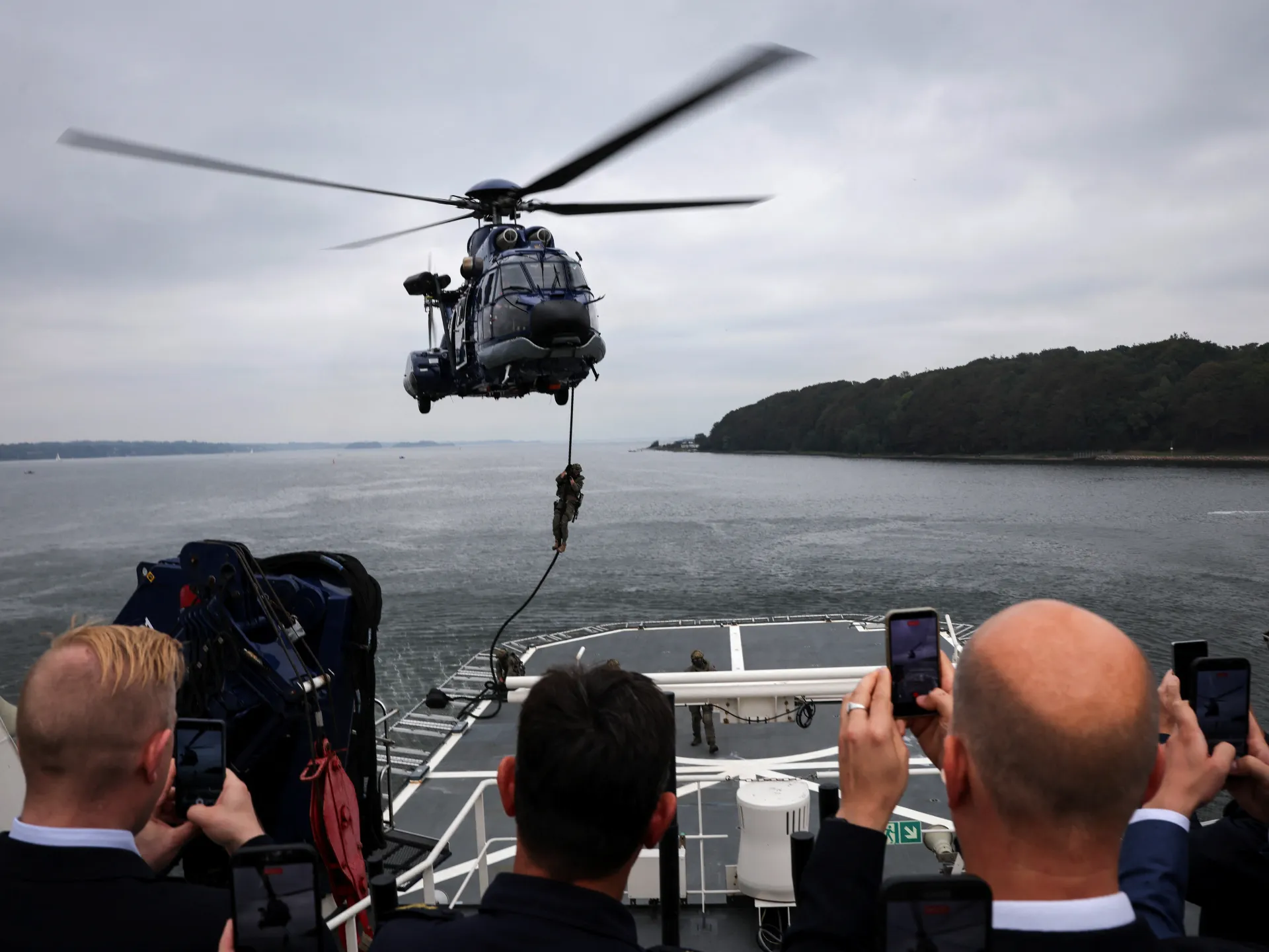 Military helicopter hoists a person above a ship’s deck while onlookers film over gray water.