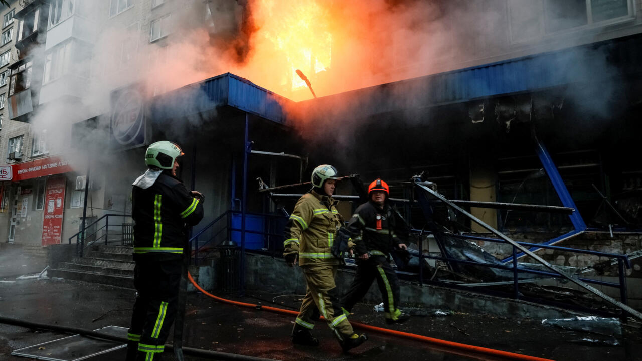 Firefighters stand outside a smoke-filled, burning storefront as flames rise above its damaged roof.