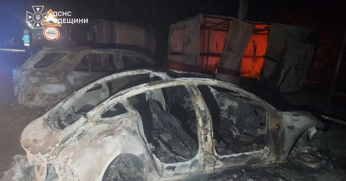 Burned-out cars near a damaged structure at night, illuminated by emergency lights.