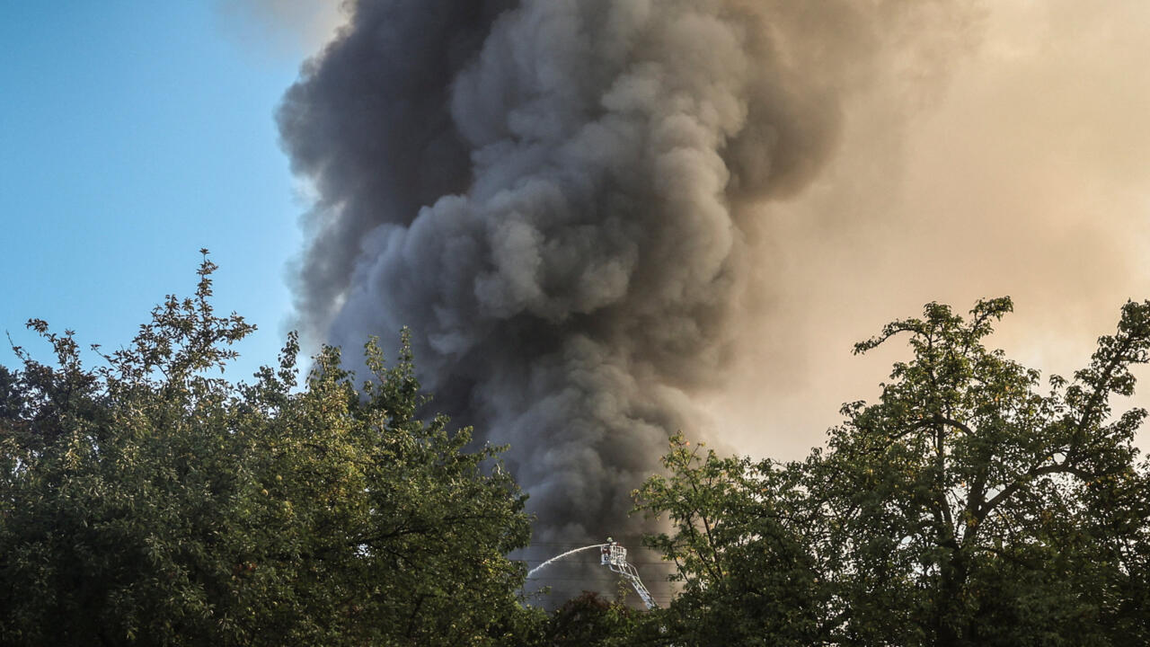 Thick black smoke rises behind trees as firefighters spray water from an elevated platform.