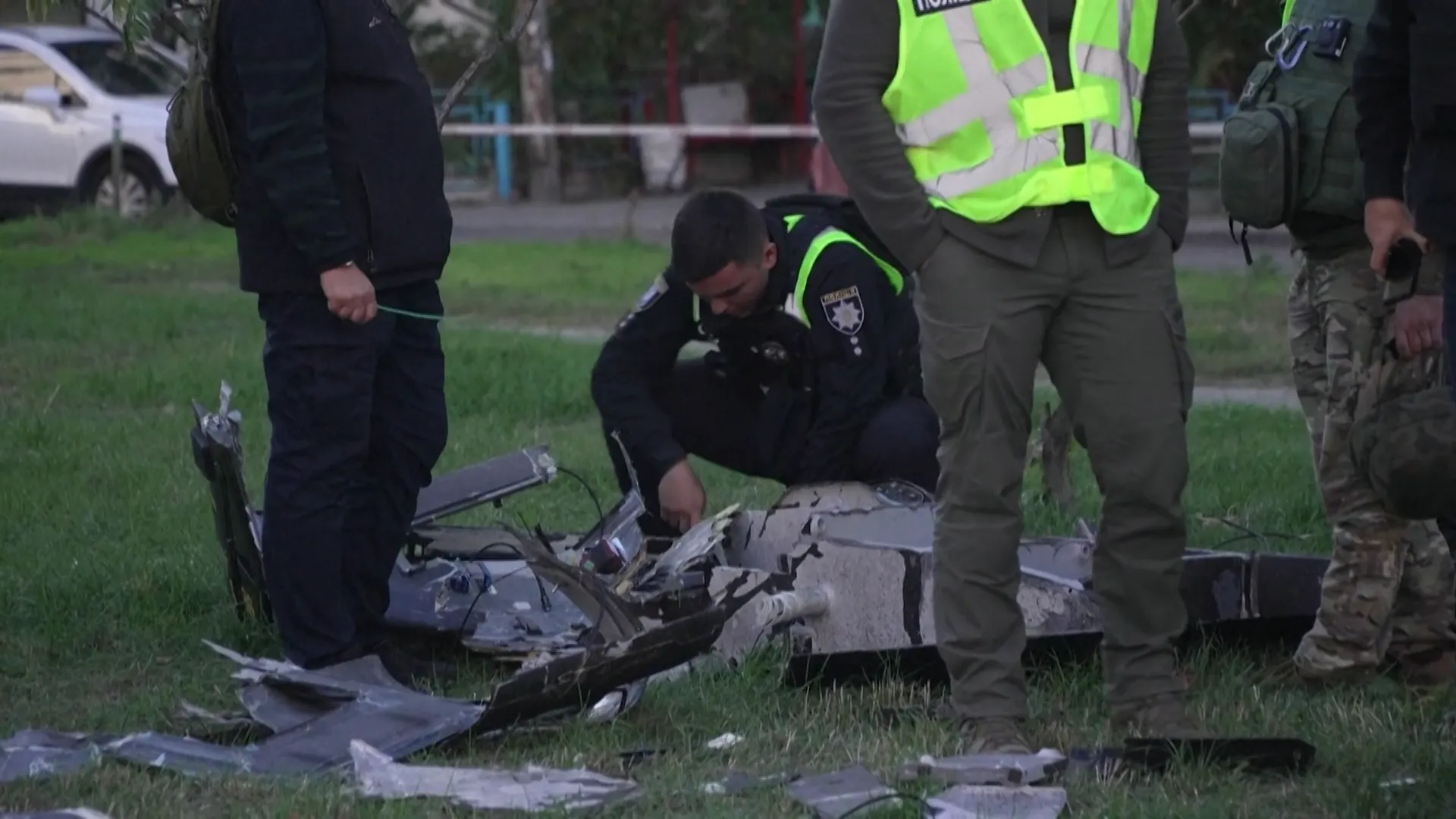Uniformed responders inspect wreckage on grass beside a road, with a police officer crouching over debris.