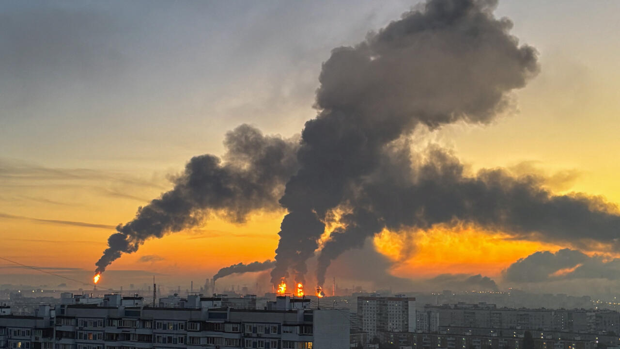 Industrial fires and towering smoke clouds rise behind apartment blocks at sunset.