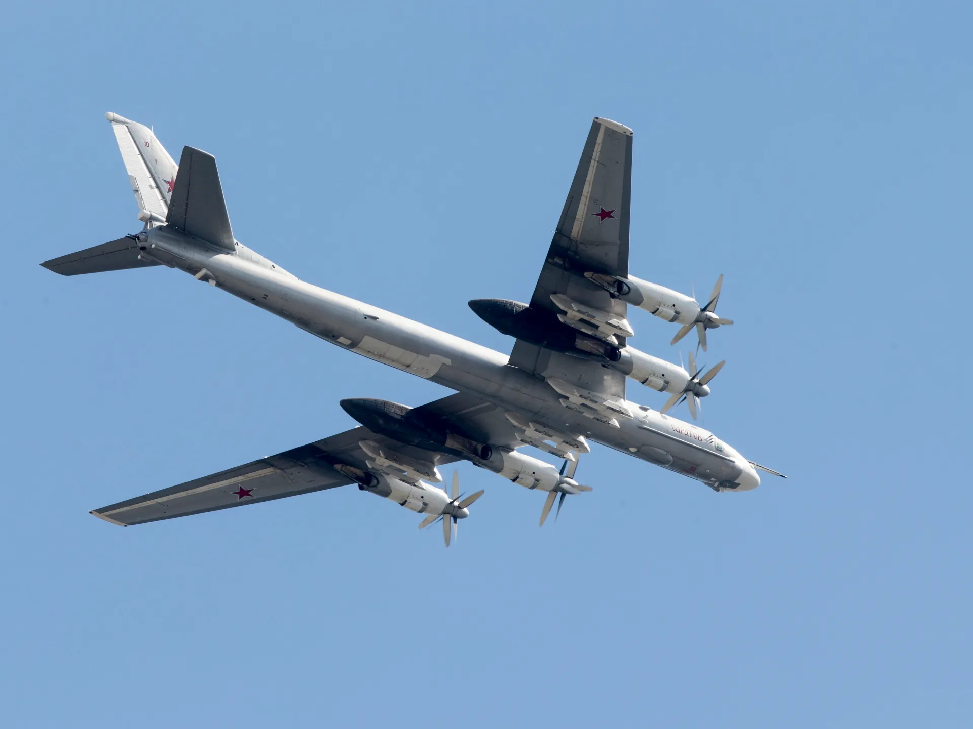 A Russian-marked four-engine propeller bomber flies overhead against a clear blue sky.