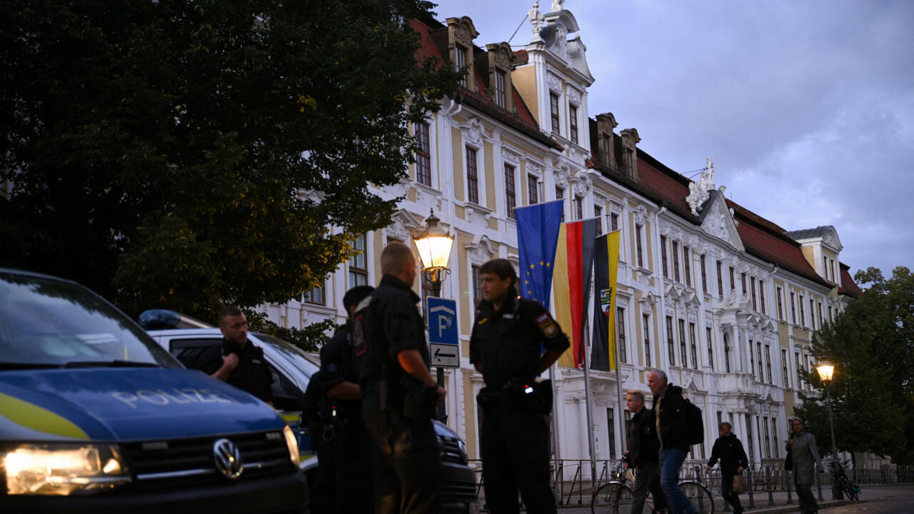 Police officers stand beside a marked police van outside an ornate building with EU and German flags.