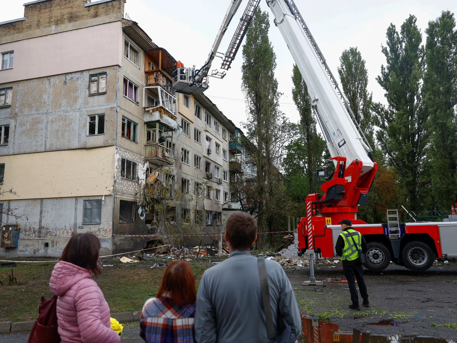 Firefighters use an aerial ladder beside a damaged apartment building as residents watch from the street.