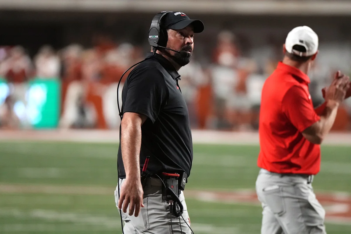 Football coach wearing a headset stands on the sideline as another staff member claps nearby.
