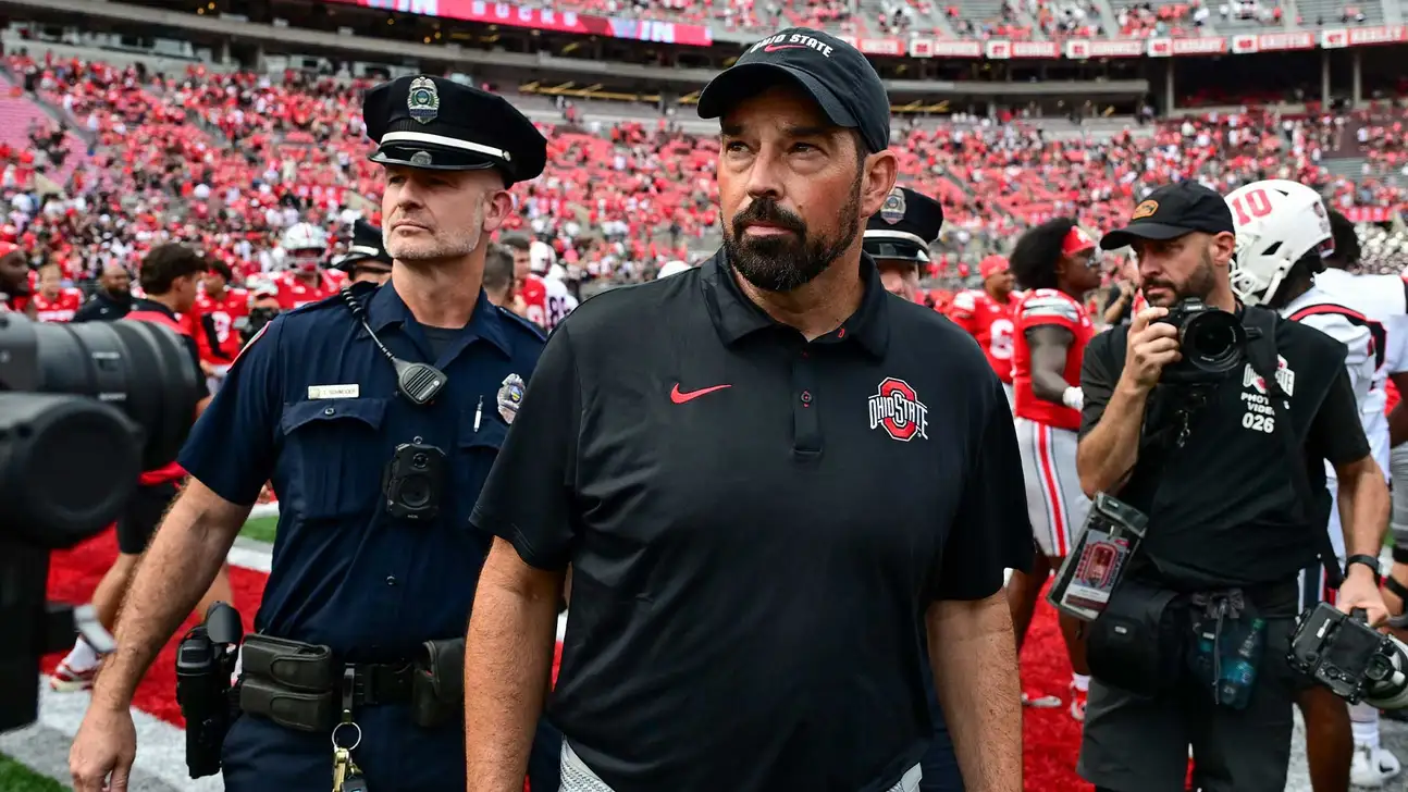 Ohio State coach in black polo walks past a police officer and photographers on football field