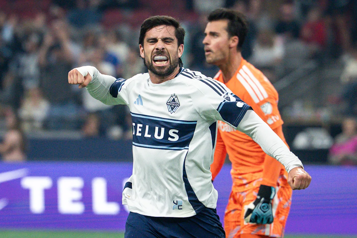 Soccer player in white jersey celebrates with clenched fists as an orange-clad goalkeeper stands behind him.