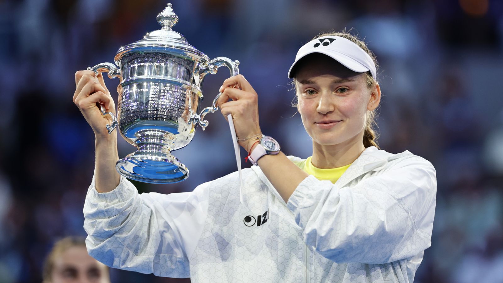 Tennis player raises a silver trophy while wearing a white visor and jacket