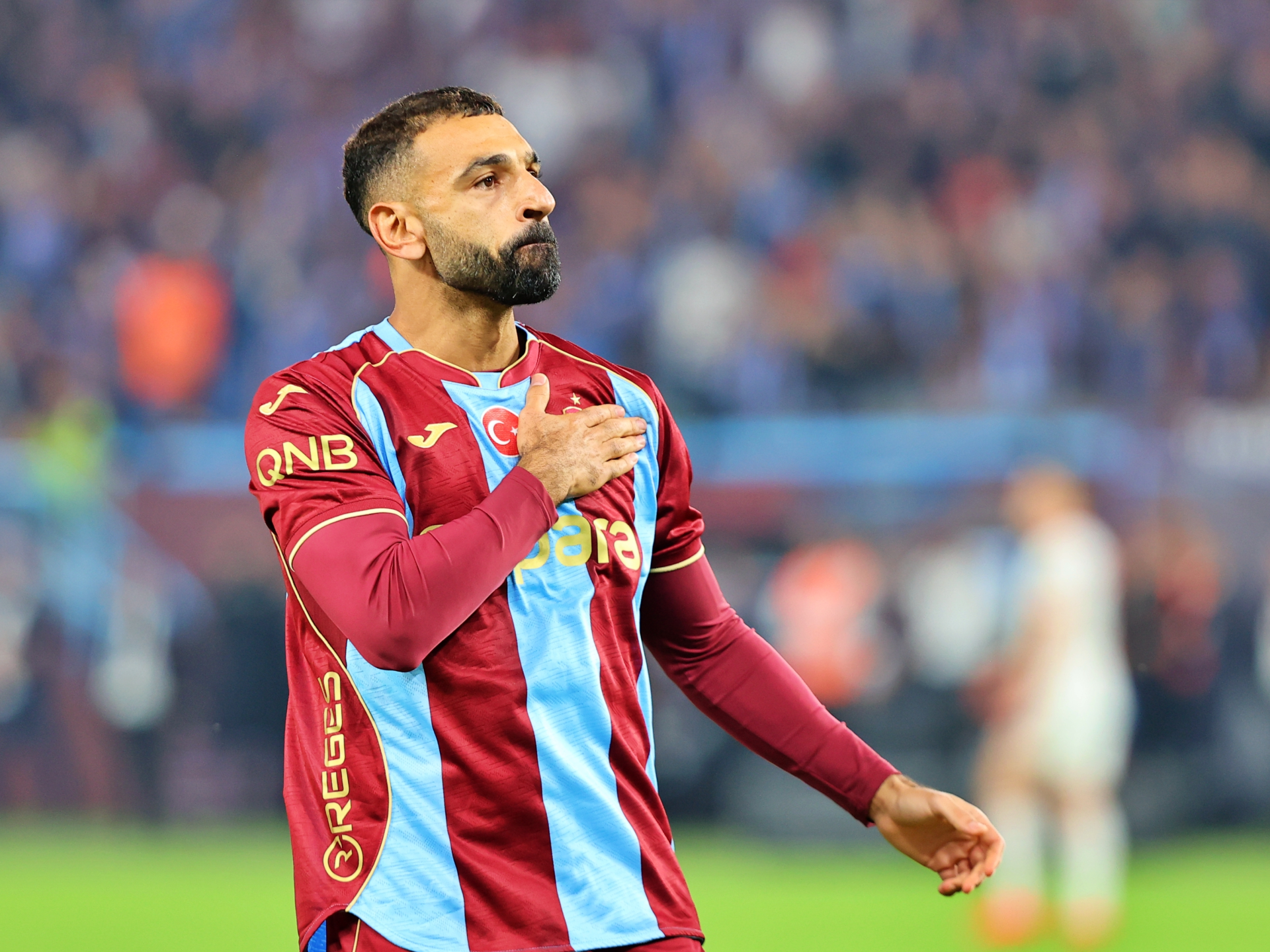 A Trabzonspor footballer stands on the pitch with one hand over his chest.