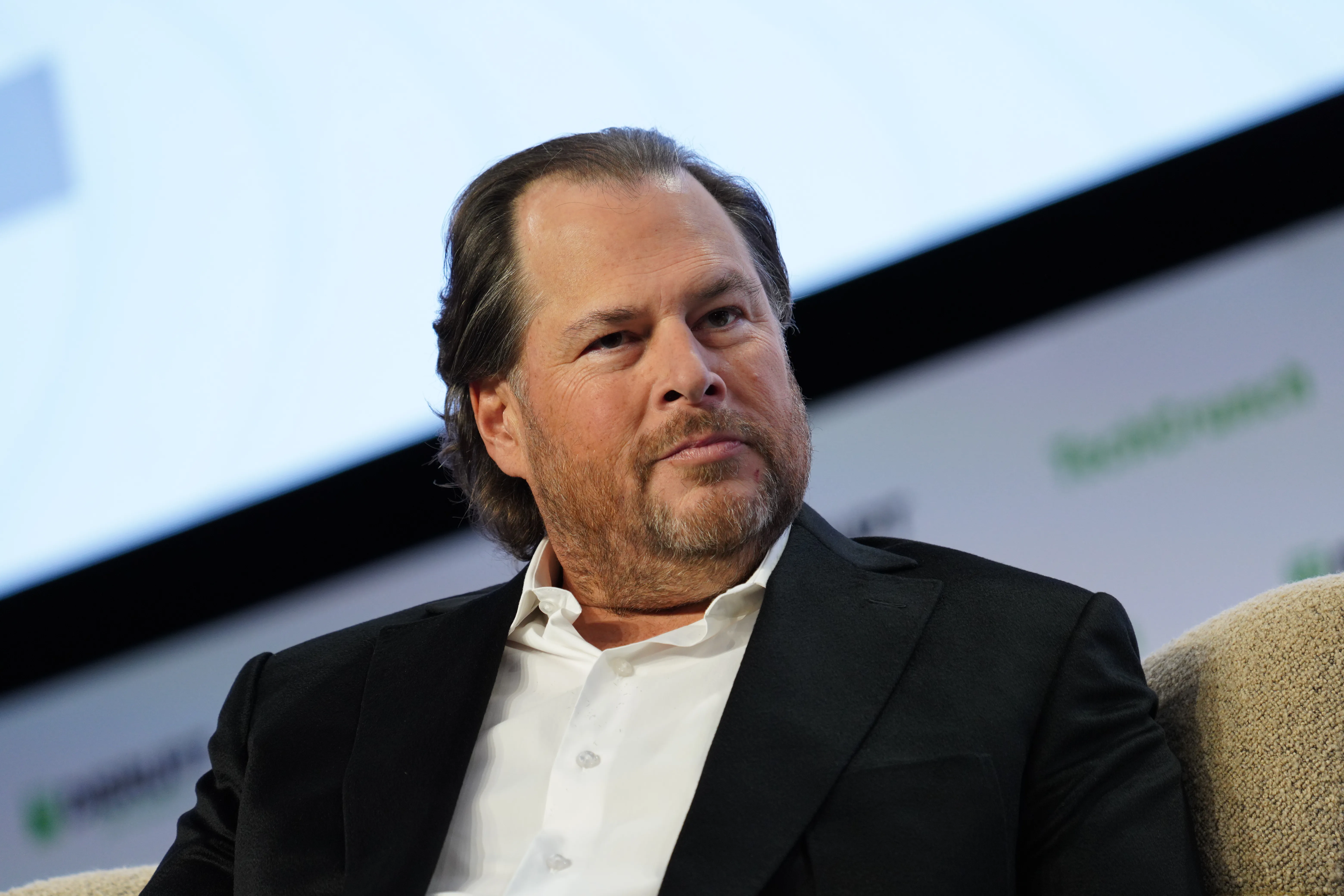 A bearded man in a black jacket sits in a chair during a staged discussion.