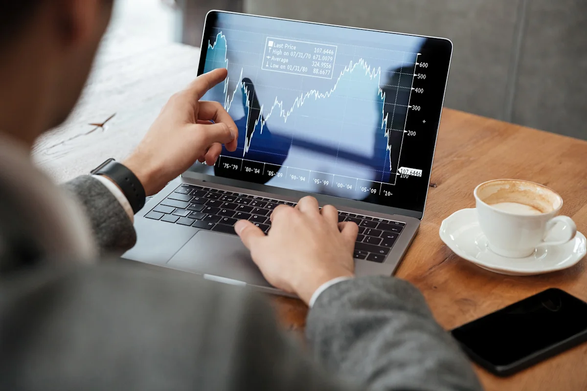 Person analyzes a financial chart on a laptop beside coffee and a smartphone.