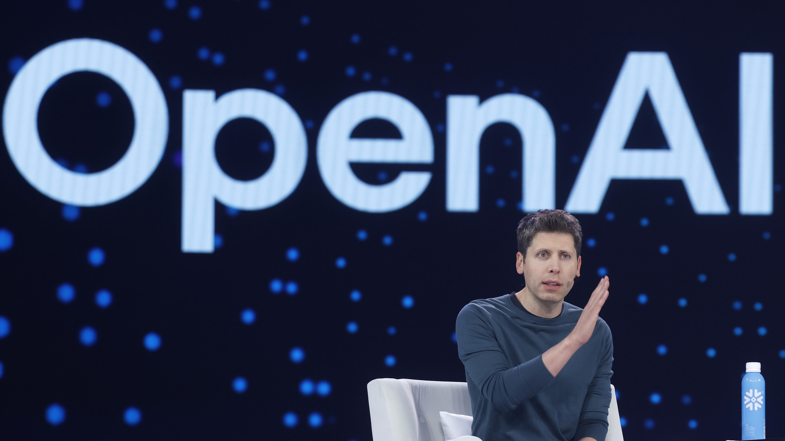 Sam Altman gestures while seated before a large OpenAI logo on a dark, starry backdrop.