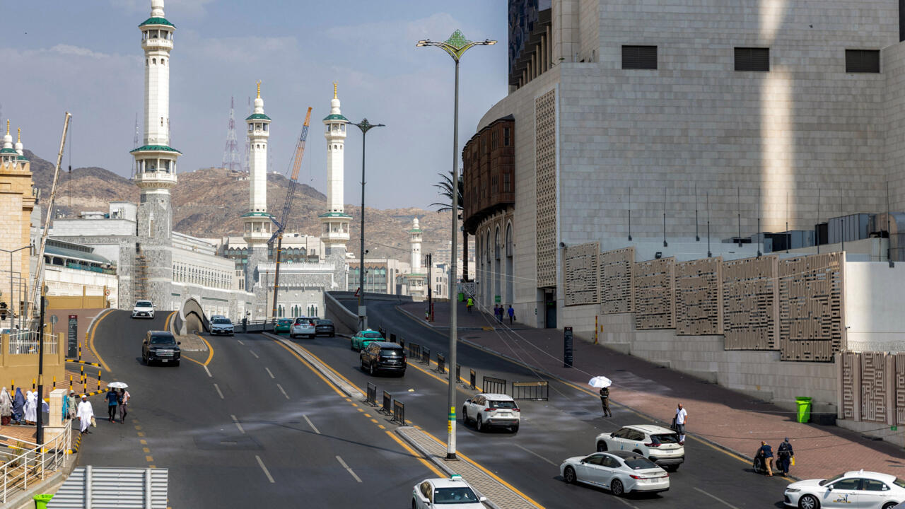 Road beside mosque complex with tall minarets, surrounding mountains, buildings, cars, and pedestrians