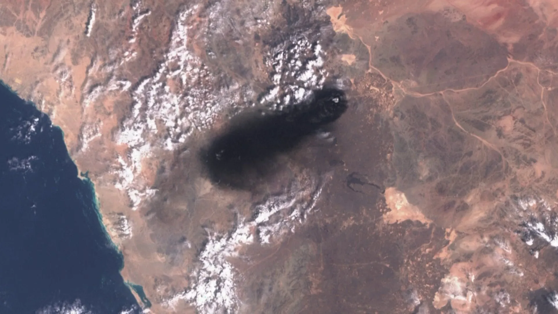 Satellite view of a desert landscape with mountain ranges, cloud cover, and a dark circular shadow