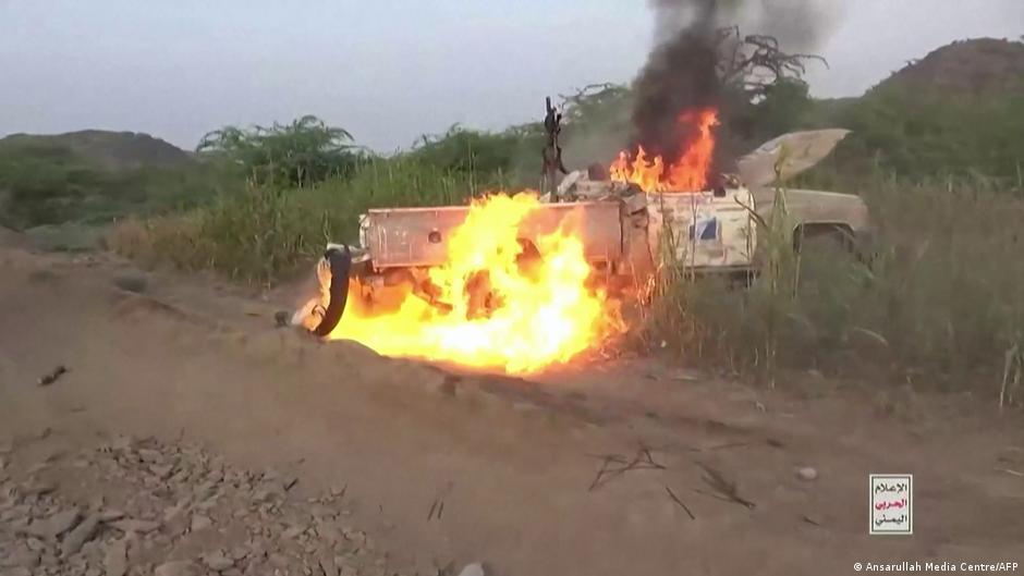 A military-style truck burns intensely beside a dirt road, sending thick black smoke skyward.