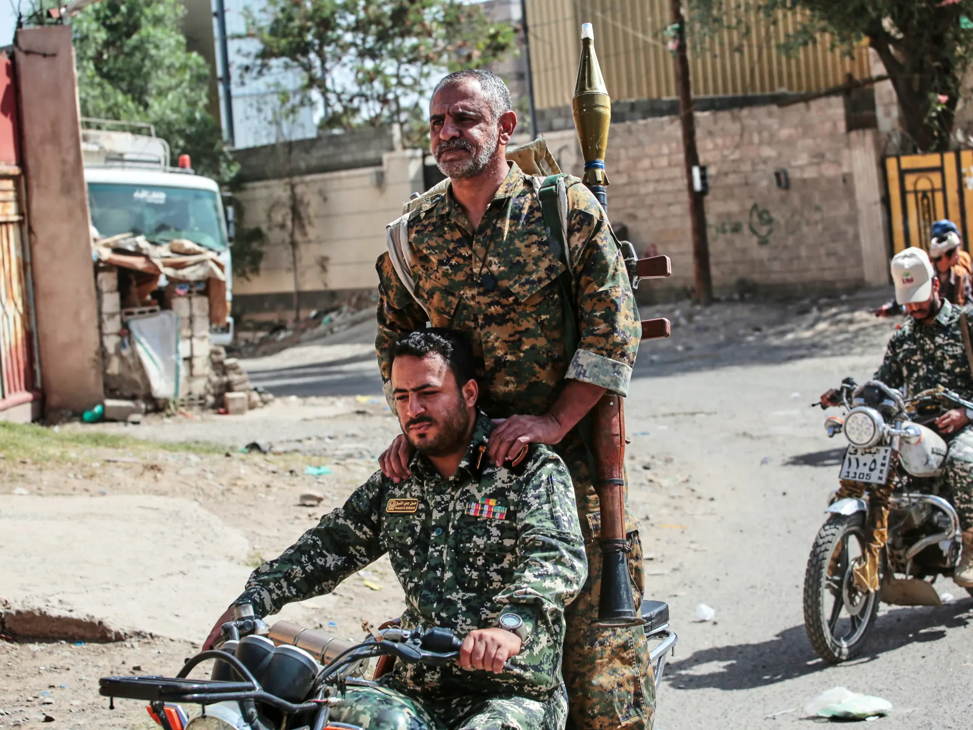 Armed men in camouflage ride motorcycles through a sunlit urban street, one carrying a shoulder-fired weapon.
