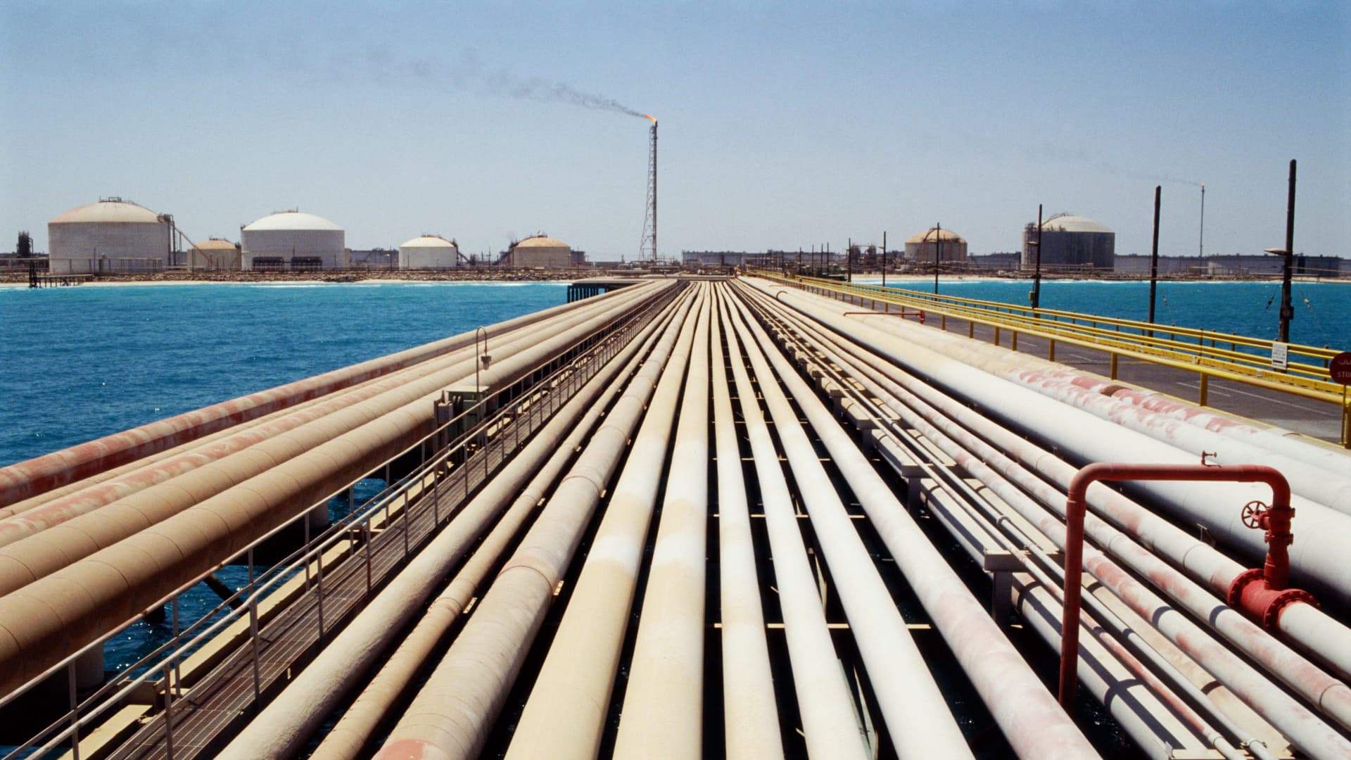 Parallel oil pipelines cross water toward storage tanks and a smoking industrial flare.