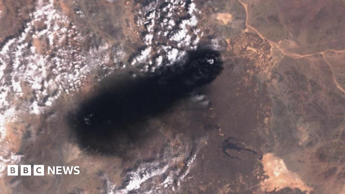 Satellite view of a large dark plume spreading across an arid landscape with scattered clouds
