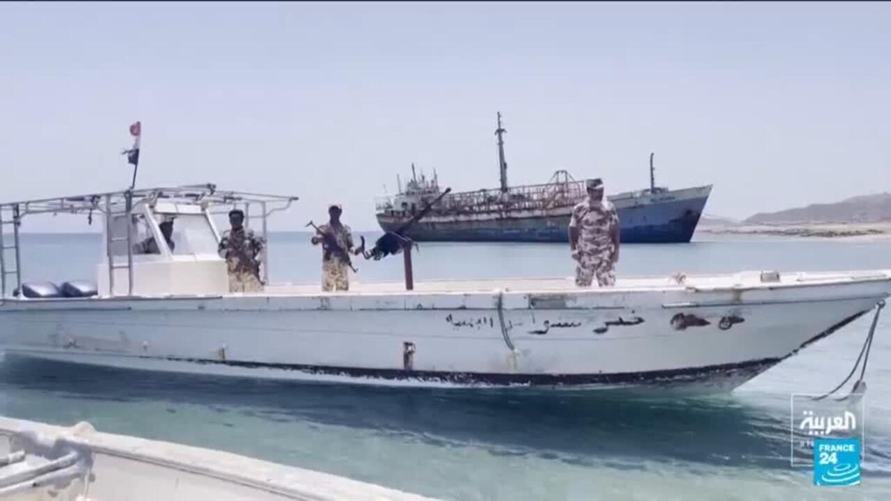 Armed men stand aboard a white patrol boat near a larger ship in calm coastal waters.