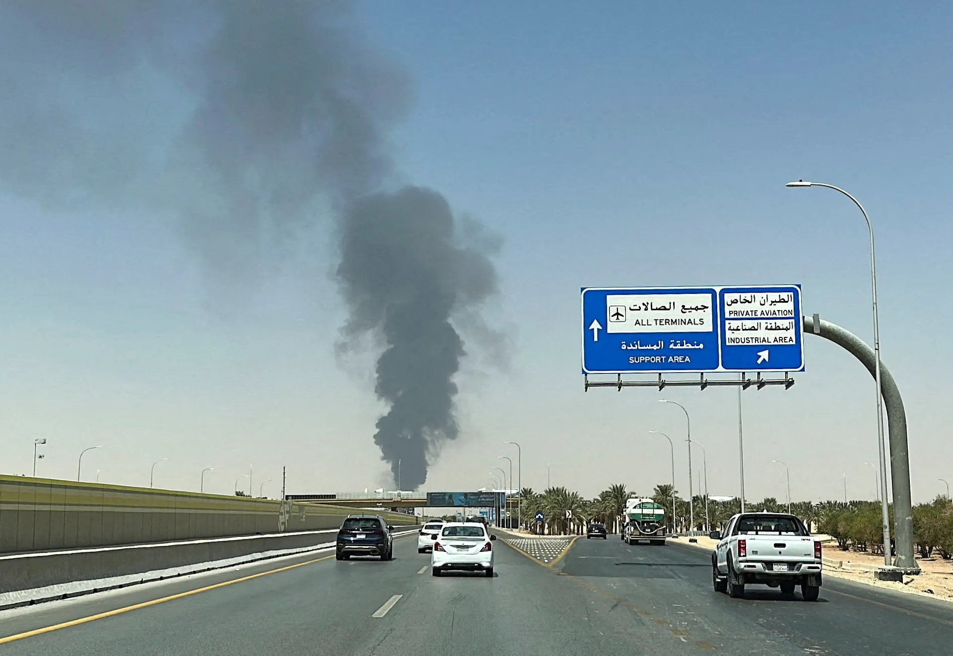 Dark smoke plume rises behind a highway with airport and industrial area direction signs