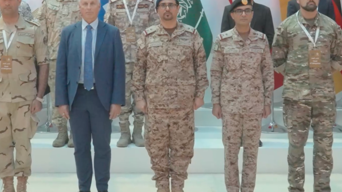Military personnel and a suited man stand in formation before national flags.