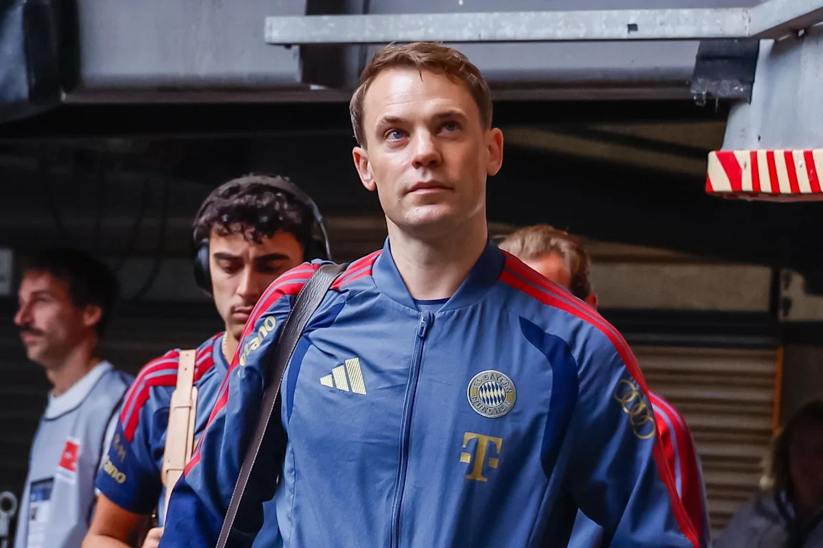 A footballer in a Bayern Munich tracksuit walks through a stadium passage with team staff.
