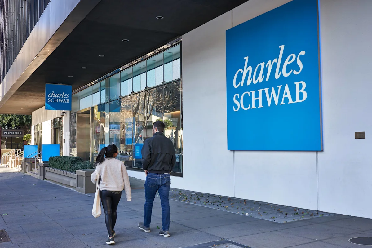 People walk past a Charles Schwab office entrance with prominent blue exterior signage.